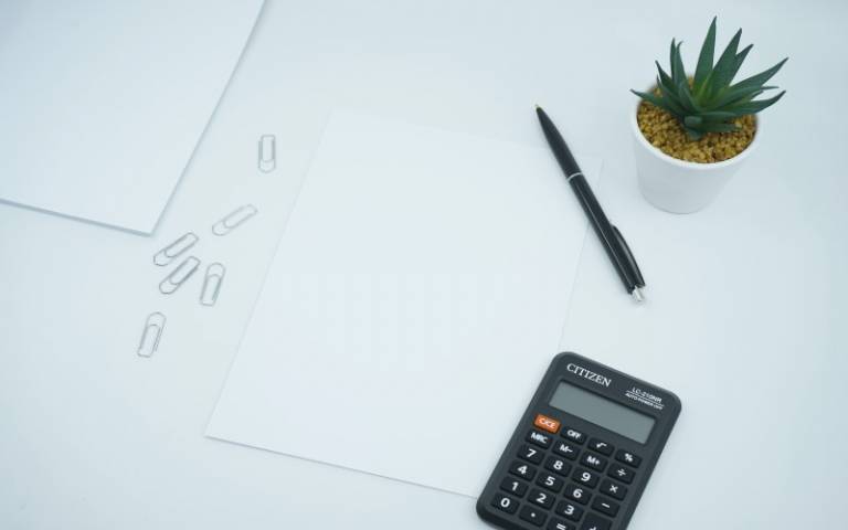 A financial themed photo featuring a blank paper on a desk next to a pen and calculator and plant
