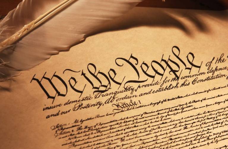 Image of a sheet of parchment and a quill; written on the parchment in Gothic font is the beginning of Article 1 of the constitution of the United States, starting with the phrase 'We the People'