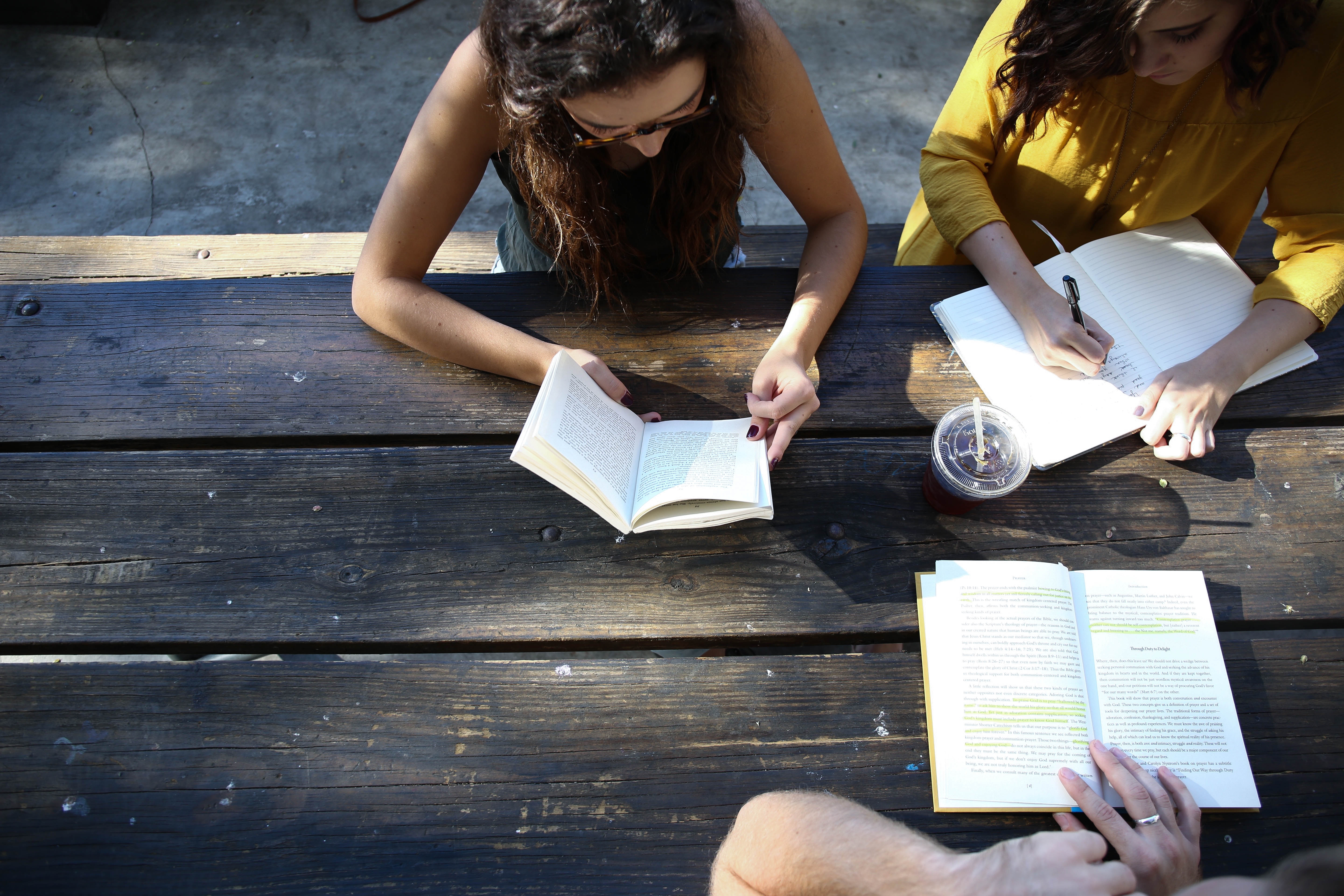Image of three undergraduate students, two females, one male, reading and taking notes around a rustic wooden table outdoors