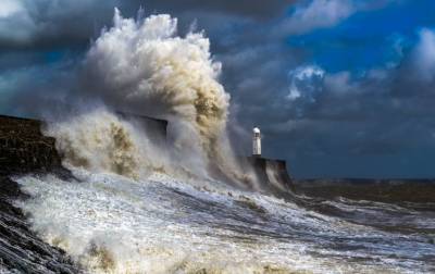 A huge wave breaks on a sea wall next to a lighthouse, throwing spray far up into the air.