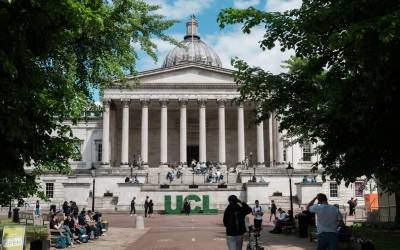 UCL's Quad in summer, the Portico building is in the distance, seen through a gap in some trees either side of the entrance, and the Quad is full of people sitting on benches or walking around. Some large green 3D letters spelling out 'UCL' can be seen.