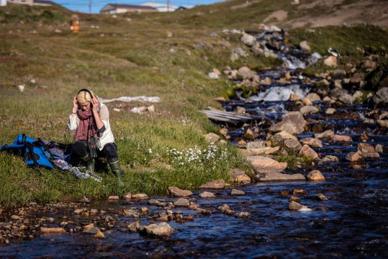 Artist Kat Austen doing fieldwork in headphones, beside water
