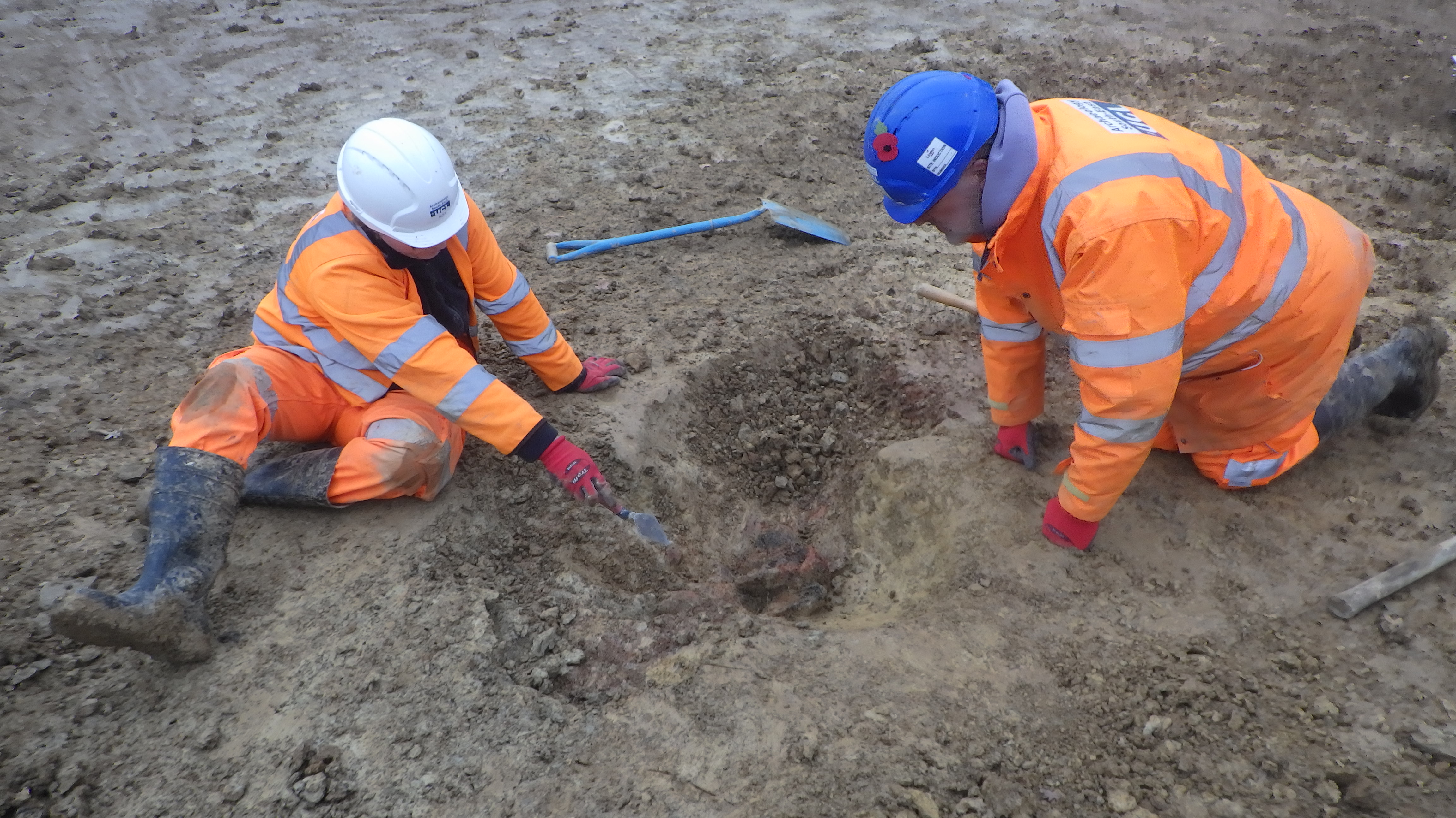 Two men wearing high vis clothing and hard hats sit on the muddy ground looking into a just-dug hole. Alex, on the left, points with his trowel at something reddish in the hole.