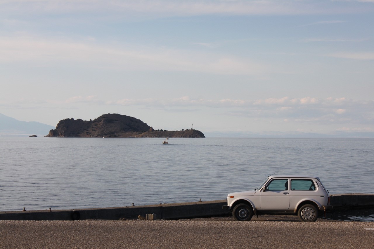 A silver car is parked in front of a large lake. There is a small island in the distance.