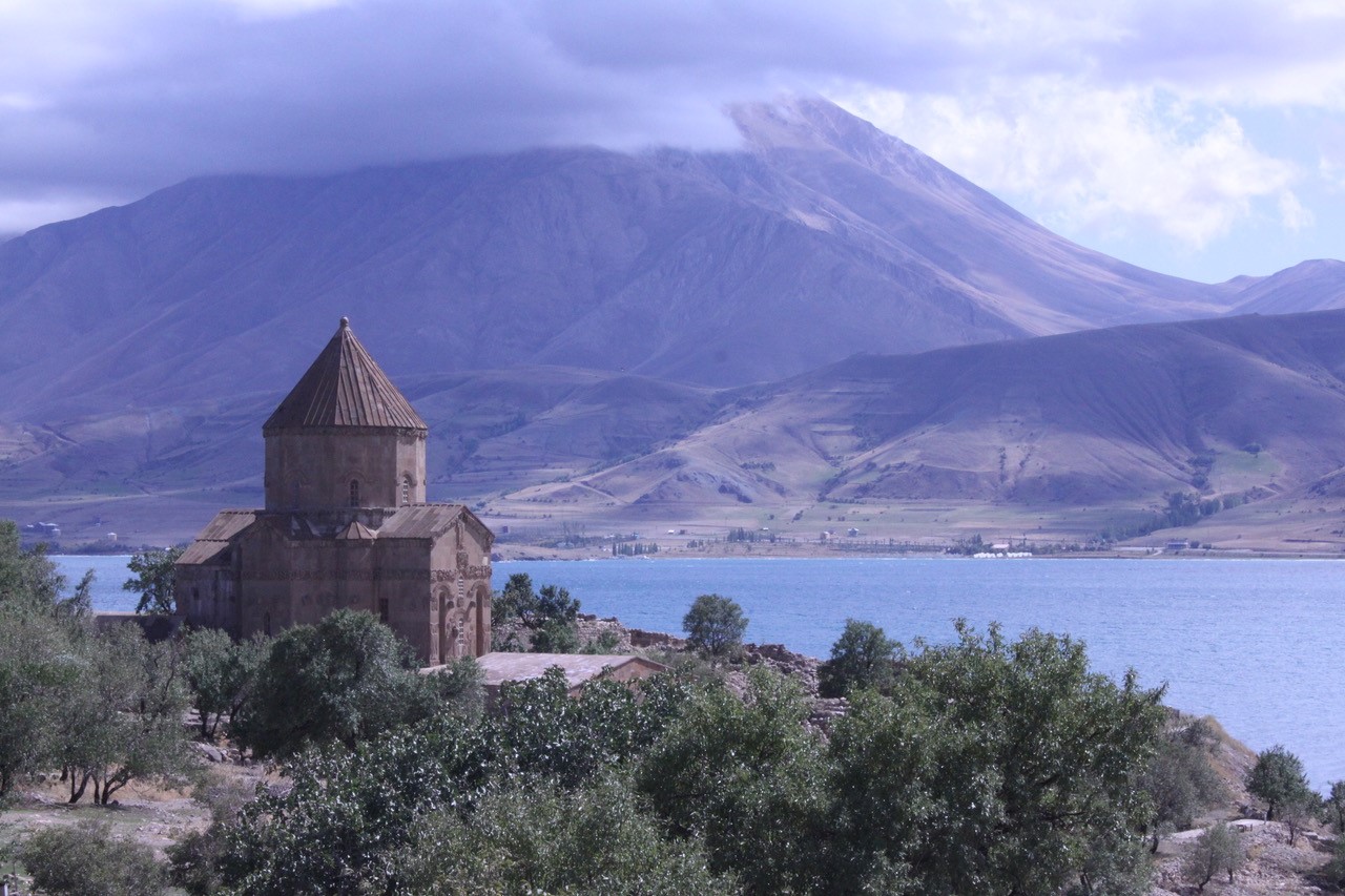 A church is surrounded by trees, with a large body of water separating it from a tall mountain.