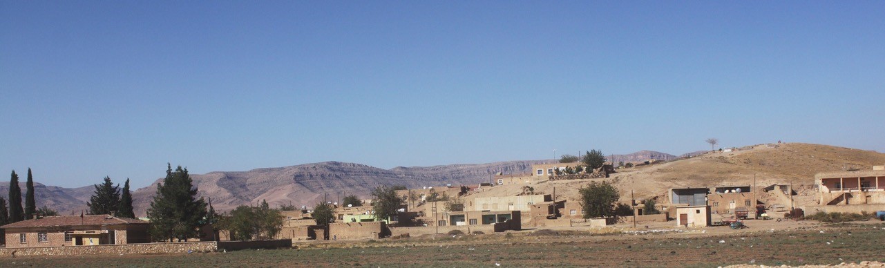 Many small buildings sit in front of a large hill. There is an agricultural field in front of the buildings. 