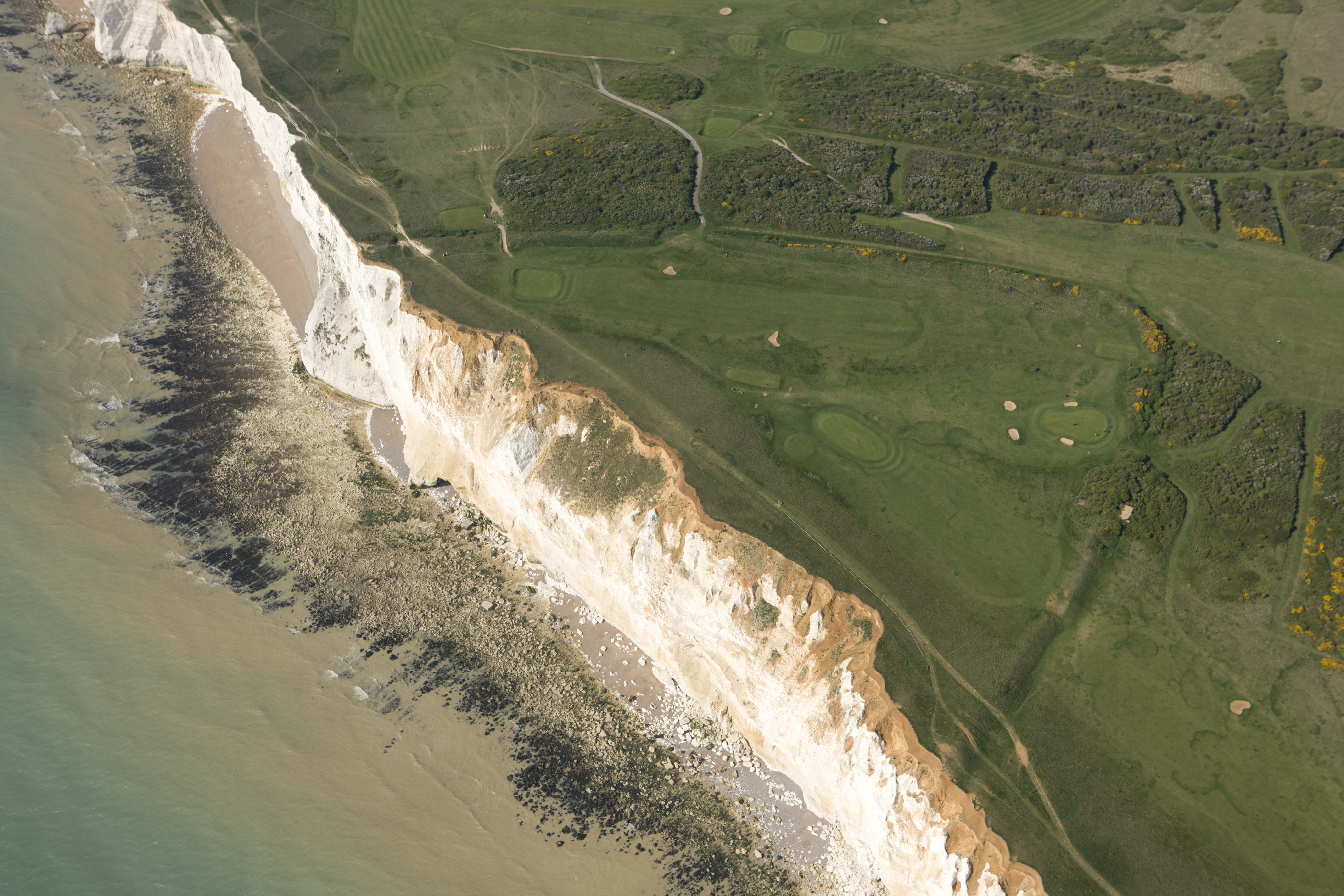 Aerial view of Seaford Head. The green land is seperated from the creamy blue sea by white chalk. Atop the cliffs can be seen a partial triangle of earthworks around the greens and bunkers of the golf course.