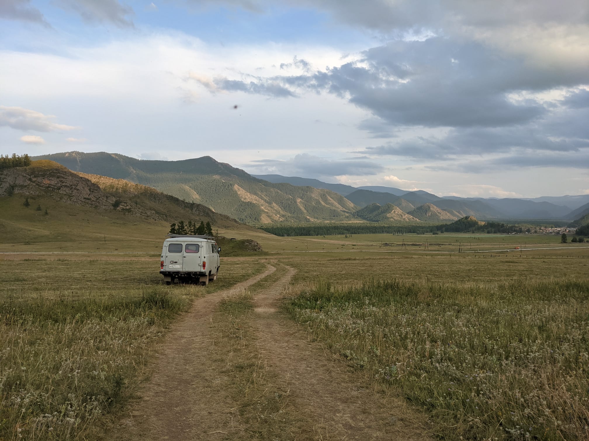 A grey van rides offroad through a field. In the distance there are layers of hills which are illuminated by the sun.