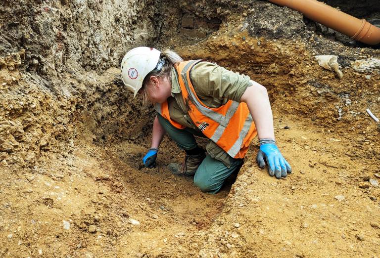 An archaeologist crouches in an excavated ditch, using a trowel to scrape at the soil in front of them.