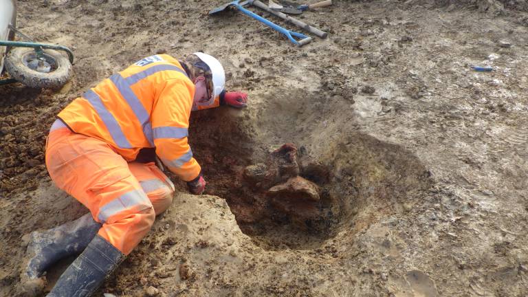 Alex supports himself on an elbow as he excavates a pit full of reddish briquetage. He is wearing high visibility clothing, a hard hat, and muddy welly boots.