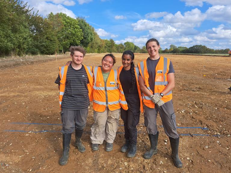Four archaeologists stand in a group, smiling. Behind them is an archaeological trench, bordered by trees. They are wearing hi-vis and wellies.