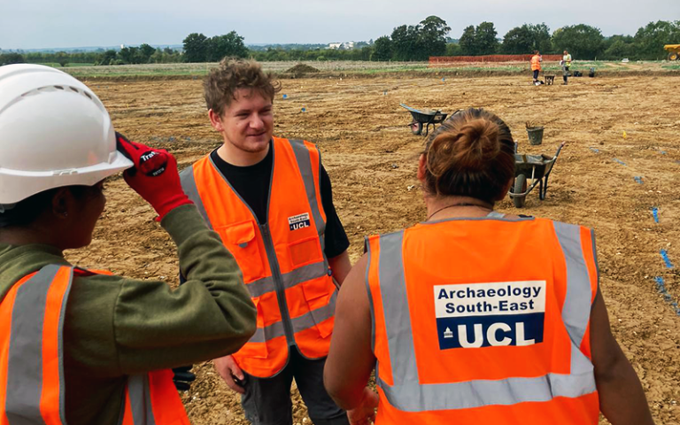 Three people wearing high visibility vests featuring the Archaeology South-East logo chat together on an archaeological site. In the background are other archaeologists and wheelbarrows.