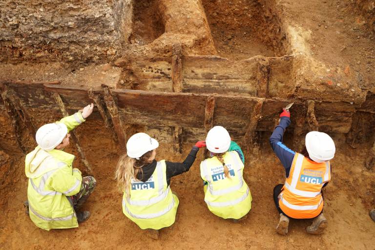 Four archaeologists sit trowling in a line. Their backs are to us and UCL Archaeology South-East is visible on their high visibility clothing.