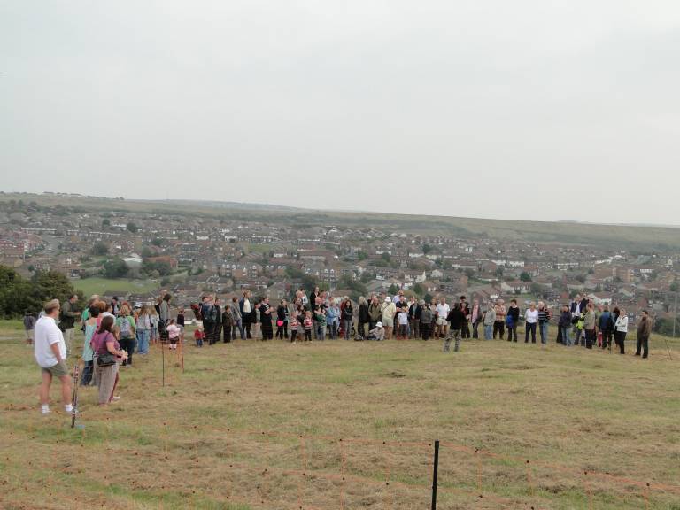 A crowd of people stand on a hill top rising above houses, listening to one person speaking.