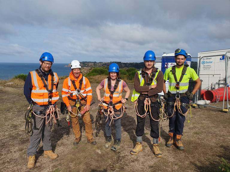 Alt text 1: 5 people stand on a clifftop with their backs to the sea. They are smiling, and wearing hardhats, hi-vis, and harnesses.