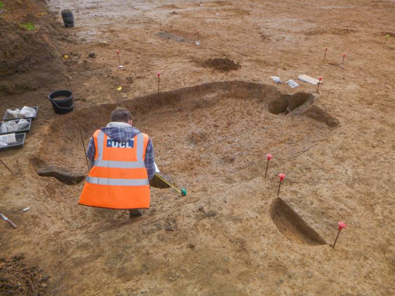 An archaeologist works inside the remains of a sunken featured building. The ovoid rectangular depression is about 4m by 2m in size, and in the middle of each short edge a posthole has been found. The archaeologist has their back to us.