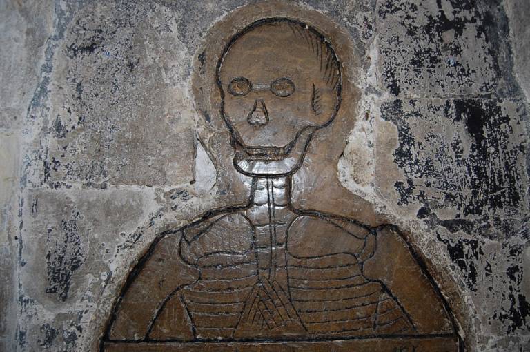 A brass engraving of a skeleton from the chest up. The skeleton’s hands are clasped on the chest.