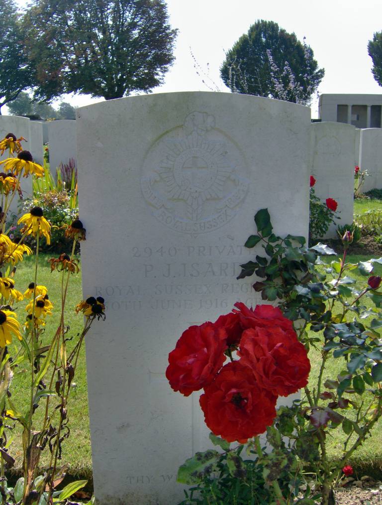 A white marble grave stone featuring the crest of The Royal Sussex Regiment above P.J. Isard’s name and date of death. The inscription is obscured by a flowering red rose.