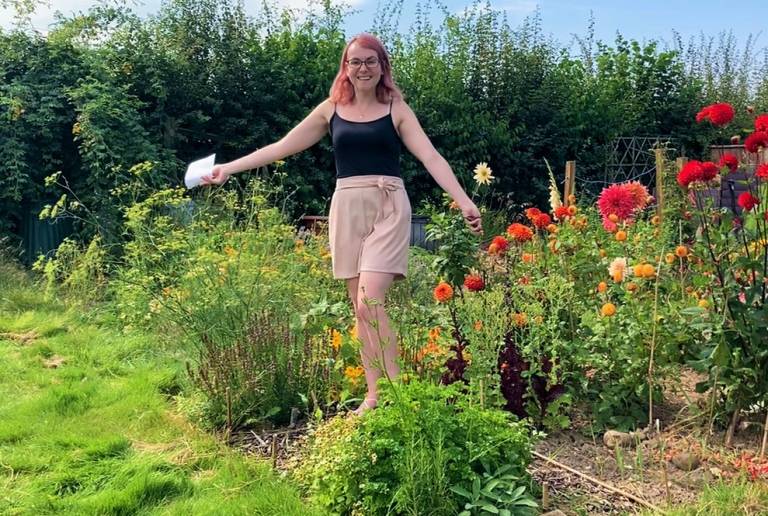 Lorna stands, arms outstretched, in her allotment. It is a sunny day and the plants are a lush green, contrasted with orange and red flowers of various heights – some nearly as tall as Lorna herself!