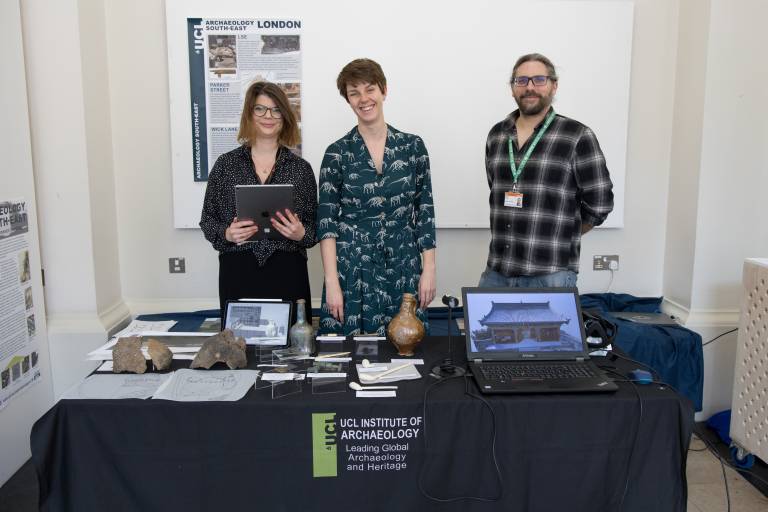 Three people stand smiling behind a table covered in archaeological finds, including clay tobacco pipes, glass vessels and large pottery fragments. A laptop displays a 3D model of a Chinese temple.