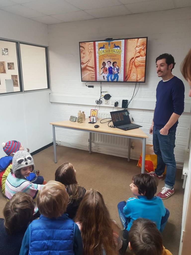 A group of young people sit on the floor. In front of them, Jake presents about his book while smiling. On a presentation behind Jake is a colourful photo of the cover of his book.  
