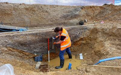 An archaeologist in a high viz jacket and wellies stands in a muddy puddle, using a rectangular tin to take a cross section of soil from the side of the excavation.