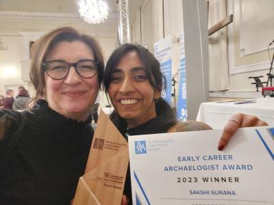 Two women grin for a selfie. Sakshi, a brown woman, holds her award up into the camera frame while Kayt, a white woman in glasses, smiles next to her.
