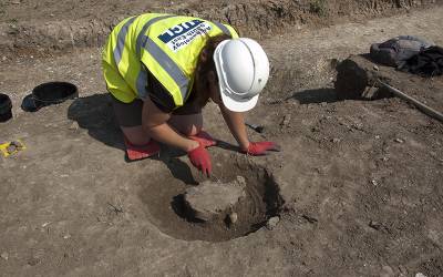 An archaeologist in a hard hat kneels down to excavate a circular feature