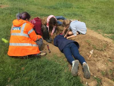 A group of children crouch by a hole in a field, digging with trowels, supervised by an archaeologist wearing orange high viz
