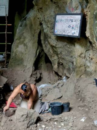A woman kneels on the ground, under a cave, digging with a trowel.