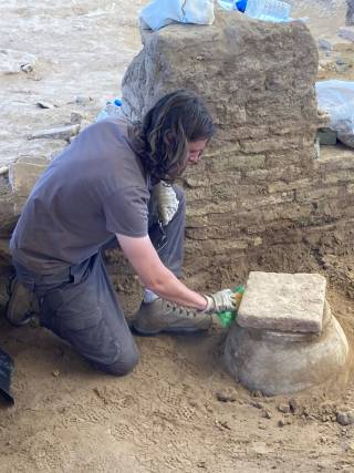 An archaeologist crouches on the ground, excavating a ceramic vessel. Behind them is a ruined wall, with archaeological equipment on it.