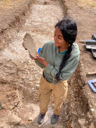 An archaeologist stands in an excavation trench, holding an artefact and smiling at it.