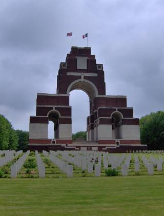 A tall, imposing, arched war memorial flying the British and French flags. In the foreground, hundreds of white headstones are arranged in neat rows.