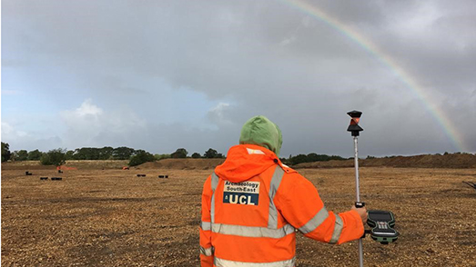 A man wearing high vis clothing stands looking at a rainbow, with his back to us
