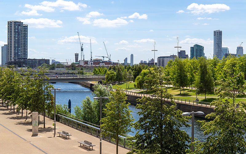 Urban green space with benches, a river and trees, with tall buildings and cranes in the background