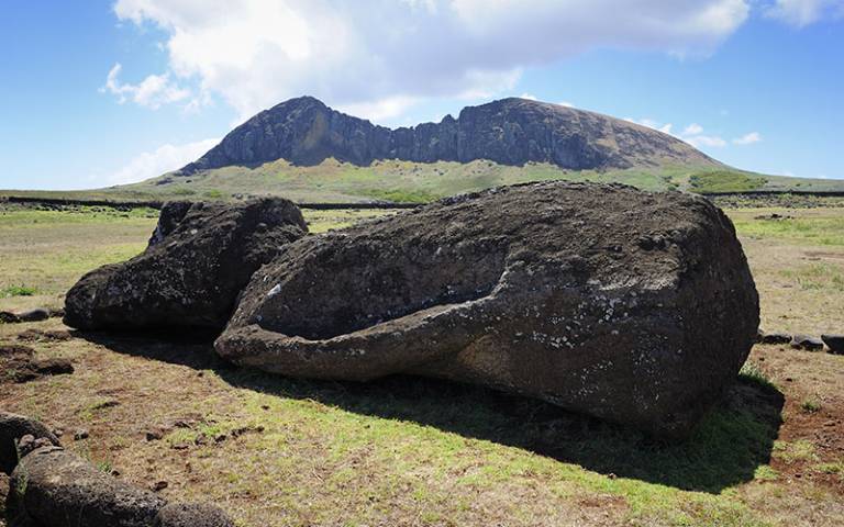 Megalithic stone statue lying on teh ground on its back with a mountain in the distance and blue sky overhead