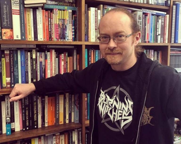 Bearded man, wearing a dark t-short and jacket, with glasses leaning against a bookshelf