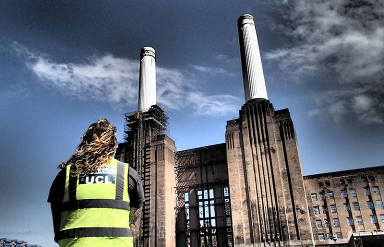 Person with long hair wearing a yellow high-vis jacket with the UCL Archaeology South-East logo on it looking at Battersea Power Station with a threatening cloudy sky overhead