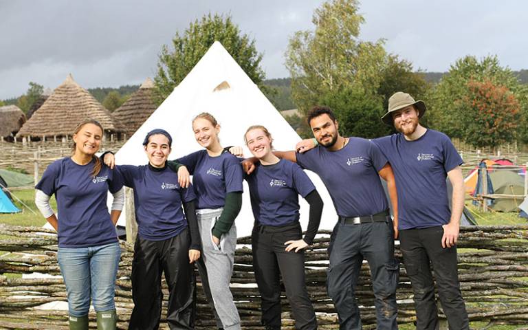 Six people (students) in branded blue t-shirts on an ancient farm with replica roundhouses in the background and camping tents in the foreground