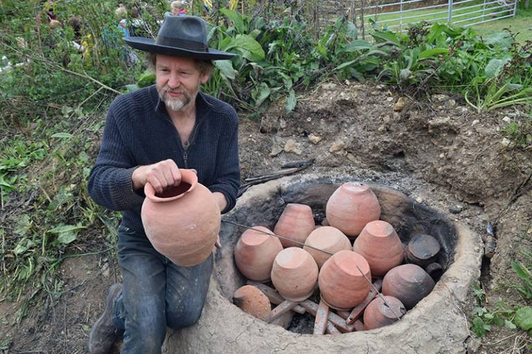 A bearded man (wearing a black top, jeans and a hat) sitting on the ground next to a pottery kiln holding a ceramic pot 