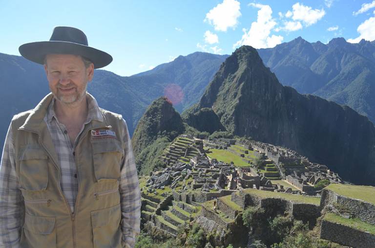 Bearded man wearing a hat, checked shirt and brown jacket, standing in a mountainous location in front of an archaeological site