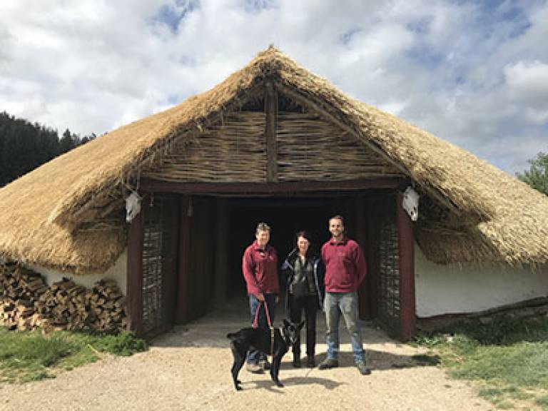 Photo of, from left to right, Maureen Page (Butser Ancient Farm Director) with her dog, Fagan; Sue Hamilton (UCL-IoA Director) and Simon Jay (Butser Ancient Farm Director) outside the farm's Iron Age round house.