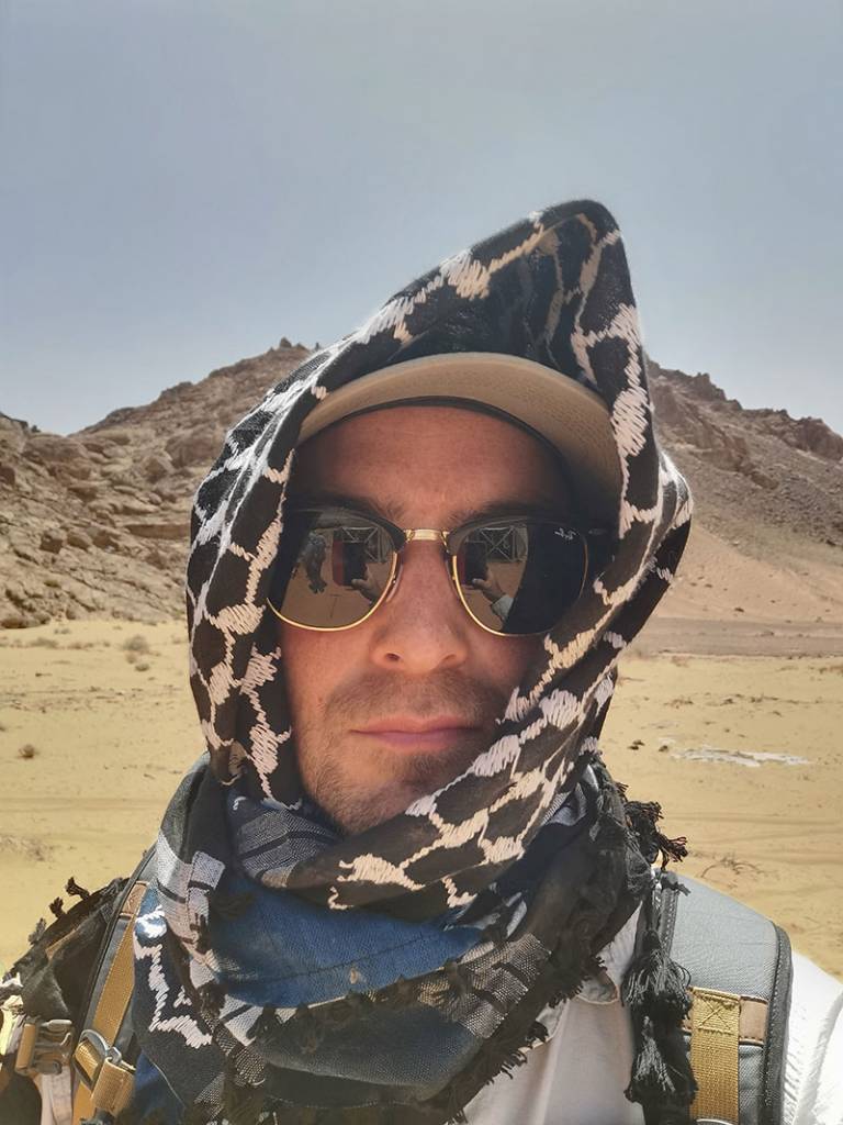 Man looking at the camera wearing sunglasses and a head covering standing in a desert location