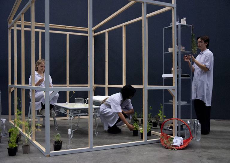 Three people, all wearing white, in and around a wooden structure (not solid) which resembles a room, with a table, seating, plants and shelving