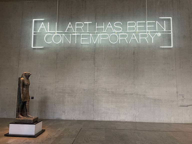 An Egyptian-deity type statue/figure in an empty grey space (gallery space?) with a neon sign hanging form the wall saying 'All Art has been Contemporary'