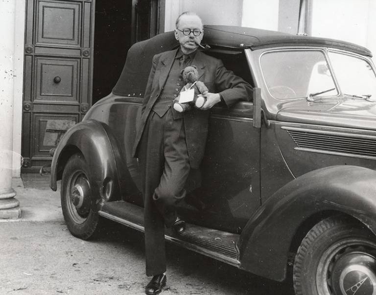 A black & white image of a man in a suit wearing glasses and smoking a pipe, holding a teddy bear, and leaning against an old-style car 
