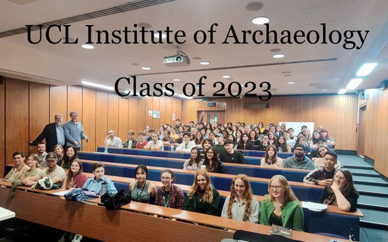 A group of students and staff sitting in a lecture theatre with black text superimposed on the image 'UCL Institute of Archaeology Class of 2023'
