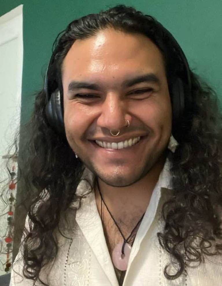 Close up of a man with long dark hair in a white top smiling at the camera, sitting in a room with green walls in the background