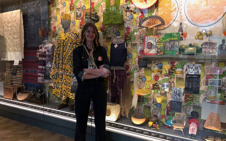 Woman with long light brown hair, wearing a black suit with a colourful shirt, standing in front of a museum display case containing multi-coloured artefacts relating to African cultural heritage