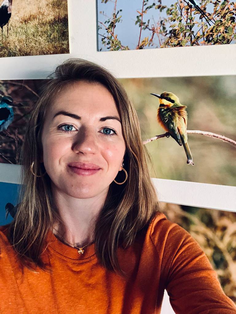 Head and shoulders picture of a woman with long light brown hair and blue eyes, wearing an orange jumper and gold jewellery, with images of wildlife on the wall beside her